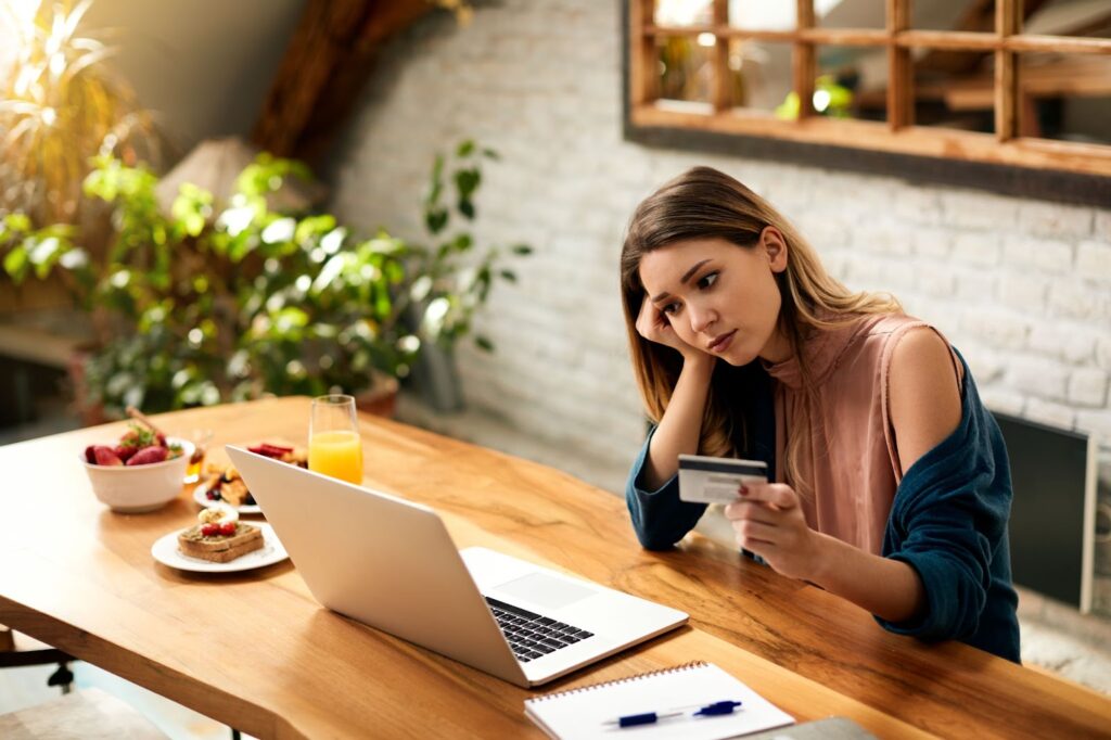 Woman feeling sad while checking her credit card over laptop at home